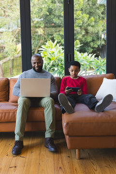 African American father and son sitting at home on leather sectional sofa using laptop and tablet