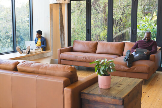 African American father and son relaxing in living room using tablet and laptop by coffee table - Powered by Adobe
