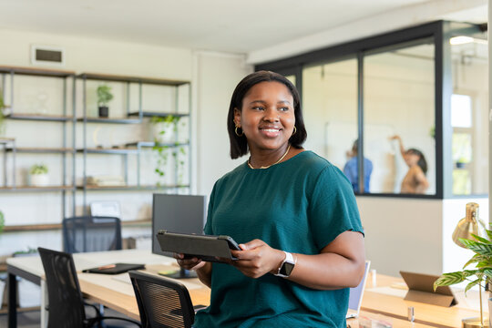 African American woman in green blouse holding tablet, reviewing screen at office desk with laptop