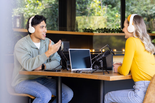Diverse coworkers recording podcast at studio table using microphones, laptop and headphones