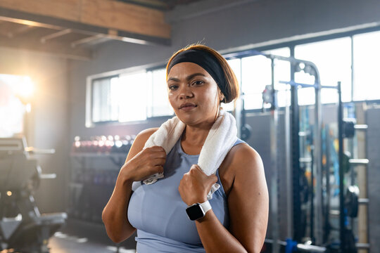 Mid adult woman standing in gym wearing light blue tank, black headband, smartwatch, holding towel