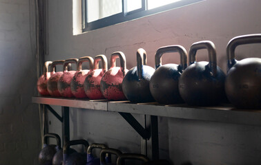 Kettlebells are sitting on metal shelf under high window, with chipped red paint and sunlit shadows