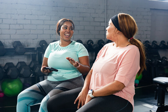 Diverse female friends wearing workout gear sitting on bench in gym holding phone near dumbbells