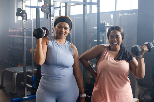 Diverse female friends wearing athletic clothing holding hex dumbbells at shoulder height at gym