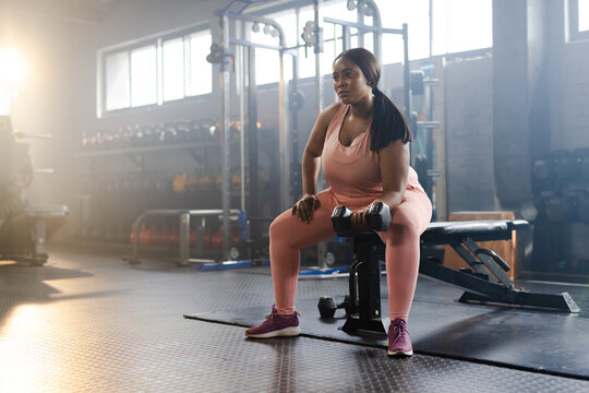 Mid-adult African American woman sitting on flat bench in gym holding dumbbells, wearing pink