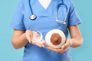 Female doctor with pink awareness ribbon and knitted breast model on blue background, closeup