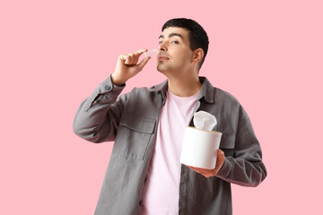 Young man with nasal drops and tissue box on pink background