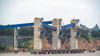 Fototapeta premium Massive industrial port terminal featuring concrete piers and large blue conveyor structures set against a forested riverbank