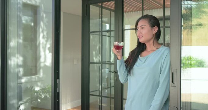 Mid adult Asian woman standing on sliding glass threshold savoring herbal tea while gazing yardward