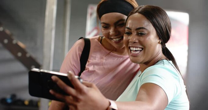 Diverse female friends holding smartphone, swiping screen, gasping at surprising result inside gym