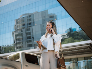 Smiling businesswoman talking on the phone and holding a tablet in front of a glass corporate building, mobile communication in the city.