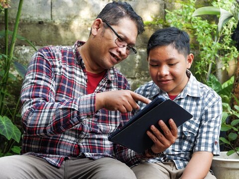 Mature Asian man in plaid shirt mentor his son using a digital tablet in a lush garden. Concept of education, fatherhood, teamwork, and remote digital learning in nature. - Powered by Adobe