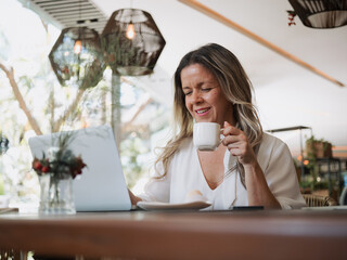 Independent woman working on her laptop and drinking coffee in a bright café, managing her online business.
