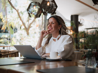 Entrepreneur smiling while thinking of new ideas in front of her laptop in a bright café, creative...