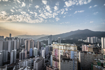 A wide-angle view of a dense urban area with high-rise buildings and green hills in the background under a partly cloudy sky.