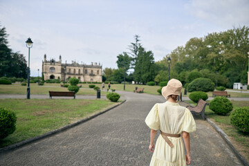 Naklejka premium woman in vintage dress walks along a cobblestone path toward a grand castle set in a manicured park, creating a serene historic ambiance
