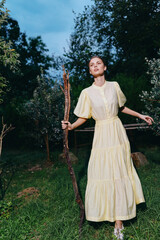 Obraz premium A woman in a pale yellow dress stands in a grassy countryside field holding a wooden staff, exuding serenity, elegance, and a graceful connection with nature