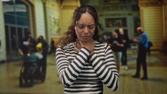 Young african american woman in a striped top holding her throat and grimacing in an ornate building concourse; choking emergency distress.