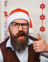Happy Ney Year. Xmas holiday celebration. Santa man at New Year show thumb up. December holiday party for man in Santa hat. Xmas man with moustache winking. Christmas shopping sale. Merry Christmas