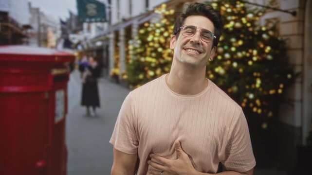 Man with hand on chest smiling and looking up, wearing glasses and a ribbed t-shirt, standing by a red postbox on a christmas decorated city street; holiday joy.