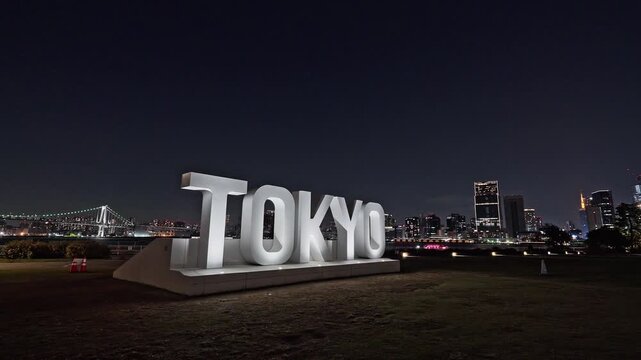 The massive, bright TOKYO sign monument stands before the glittering Tokyo skyline and Rainbow Bridge at night.
