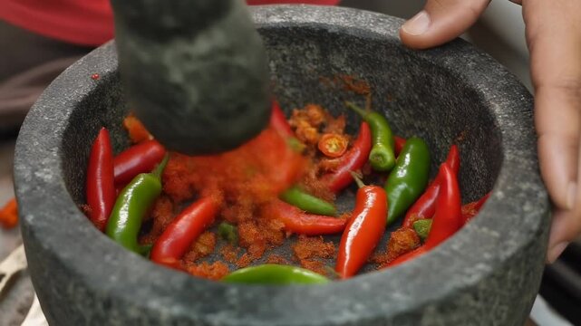 A close-up shot of a hand grinding red and green chili peppers with a pestle in a dark stone mortar, preparing a spicy paste.