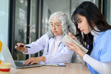 Fototapeta premium Old asian woman physician pointing at laptop showing to young doctor taking note sits at exam table.