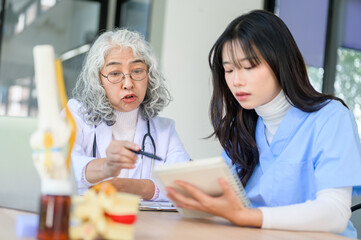 Obraz premium Old asian woman physician teaching and pointing at notebook of a young doctor sitting at exam table.