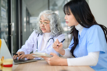 Fototapeta premium Old asian woman physician teaching and typing on laptop with young doctor taking note at exam table.