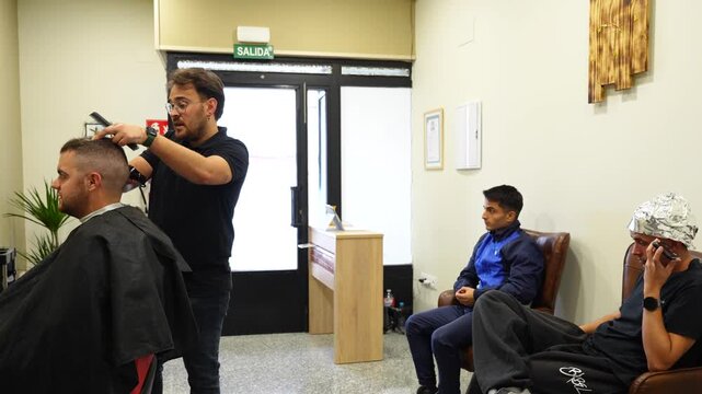 Wide view of a barber cutting a client's hair with two waiting customers seated behind, one talking on a phone