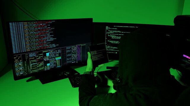 Dark hooded figure decrypts digital code using foldable screen in green-lit room