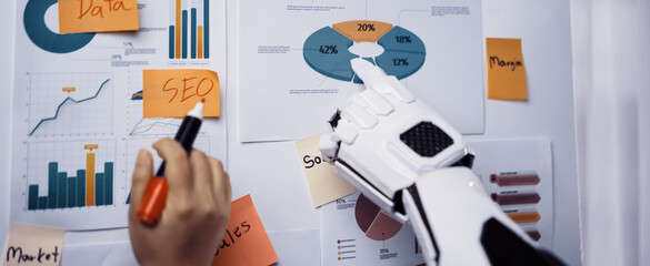 A robotic arm interacts with colorful infographics featuring business metrics, showcasing modern technology's role in data analysis and creative planning in an office setting.Noogenesis