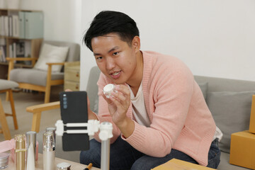 Asian young man demonstrating skincare cream jar during live product presentation with various cosmetic bottles on table, cozy living room setting, confident expression