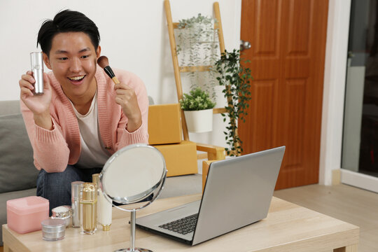 Asian Young man holding skincare bottle and makeup brush smiling at laptop mirror on table while recording beauty tutorial in cozy living room - Powered by Adobe