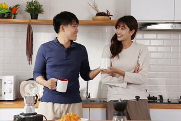 Asian young couple smiling in kitchen while sharing coffee mug and glass