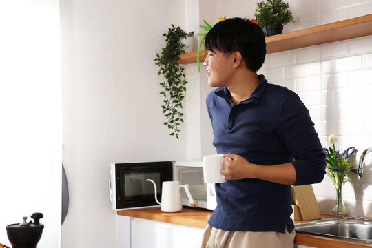 Asian Young man enjoying morning coffee by bright kitchen window, holding mug and smiling while looking outside with plants and countertop appliances visible - Powered by Adobe