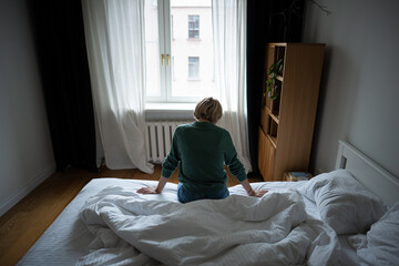 Dressed middle aged woman sits on bed in bedroom unmotivated to get up, body pose showing heavy...