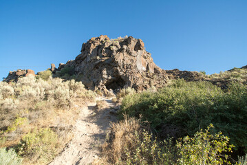Hiking trail winding through high plains scrub including sagebrush and dappled by late afternoon shade, to large rock formation under a clear blue sky. 