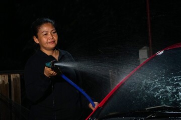 A woman is spraying water to wash a red car with a hose and nozzle against a dark, nighttime background.