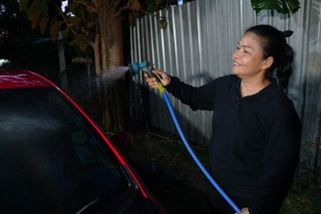 A woman is smiling while she sprays water to wash the windshield of a red car during dusk.