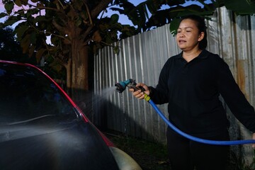 A woman is spraying water onto the hood of a red car near a corrugated metal fence line.