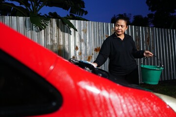 A woman is using a sponge to scrub the red car's windshield area, holding a green bucket in her hand.
