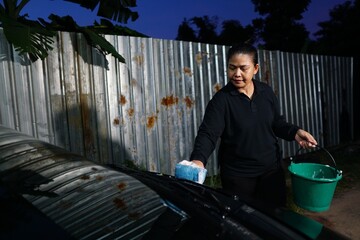 A woman is using a sponge to scrub the red car's windshield area, holding a green bucket in her hand
