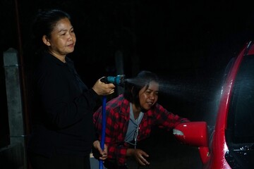 A woman is spraying water towards her friend who is bent over, washing the car's headlights.
