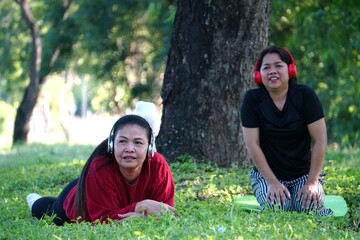A woman is lying prone on a pink mat on the grass in a resting pose, while the other woman is kneeling behind her, leaning against a tree trunk.