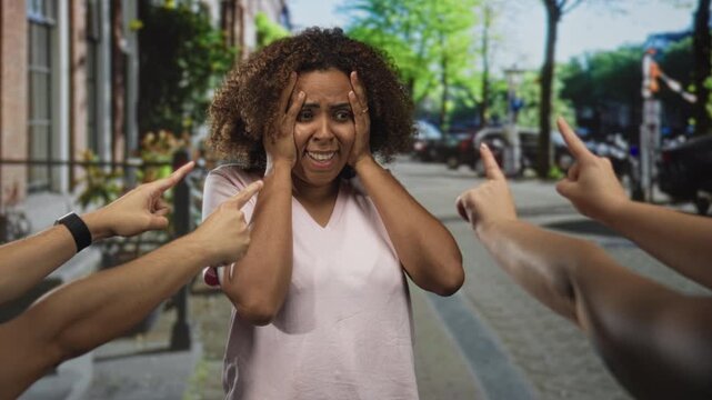 Woman with hands on head and multiple fingers pointing at her on a busy city street with parked cars; isolation anxiety.
