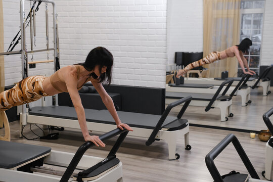 Pilates reformer workout with woman standing on carriage, using straps for controlled core training, stretching and physical therapy. - Powered by Adobe