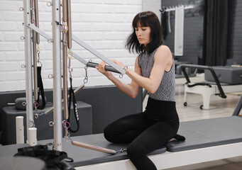 Woman preparing for pilates reformer workout by examining and adjusting the machine in modern fitness studio before exercise session begins