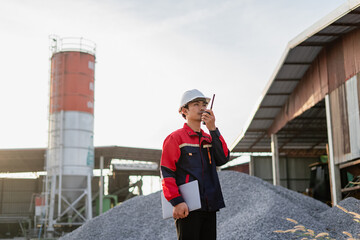 An engineer standing at industrial material storage yard checking data plans on laptop. Project management quality control. Large cement silo and pile of materials in background.
