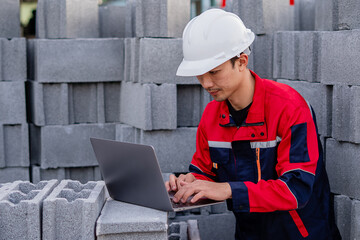 Male construction engineer wearing hard hat working on laptop using concrete blocks as a temporary workbench. Digital technology and construction process. Quality control documentation.